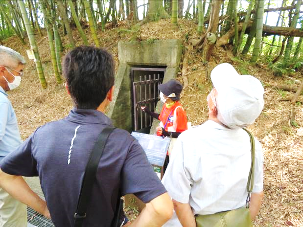 講師が、竹林にある壕の解説をしている。複数名の参加者が真剣にそれを聞いている様子。