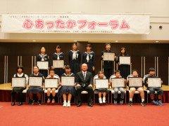スーツ姿の男性を中心に賞状を持った小中学生たちがステージの前で2列に並び記念撮影する様子の写真
