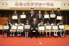 スーツ姿の男性を中心に賞状を持った小中学生たちがステージの前で2列に並び記念撮影する様子の写真