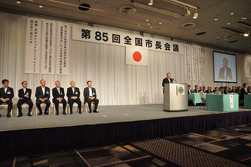 全国市長会総会でステージの上にスーツを着た人たちが椅子に腰かけて並んでいる写真