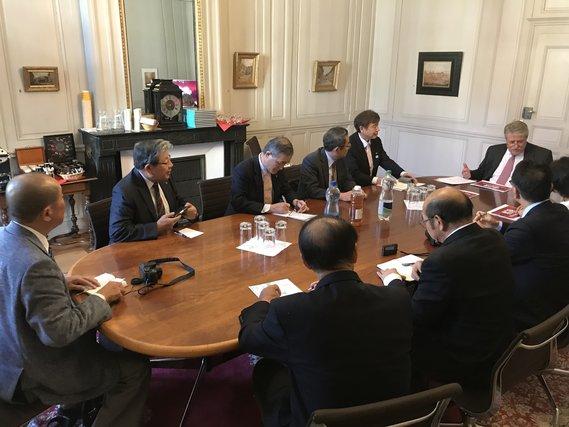 カーペットが敷かれ木製の机が置かれている部屋で着席し説明を受ける全国市長会欧州都市行政調査団の写真