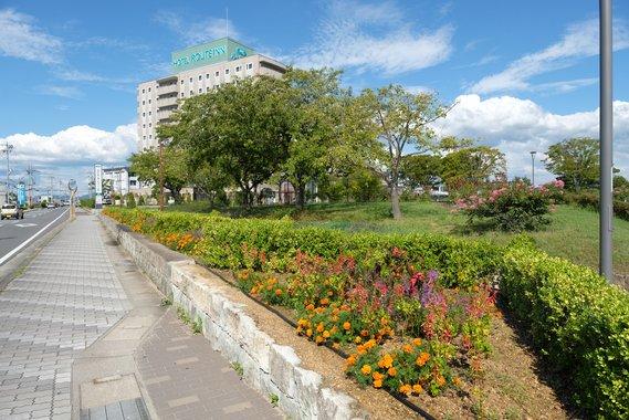 ホテルルートインを背景に、沢山の木々やカラフルな花がガーデニングされた広場の写真