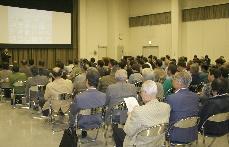 大きなスクリーンに映っているパワーポイントを座って見ている沢山の人たちの写真