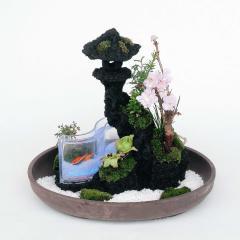 底が薄い丸い鉢に岩や植物が配置され、小さな水槽で魚が泳ぐ様子が表現されている発光ダイオード仕かけセンサリーガーデンの写真
