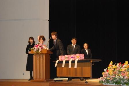 壇上のマイクの前に立つ新成人と手話の方、横に置かれた机には箱が4つ乗っている写真