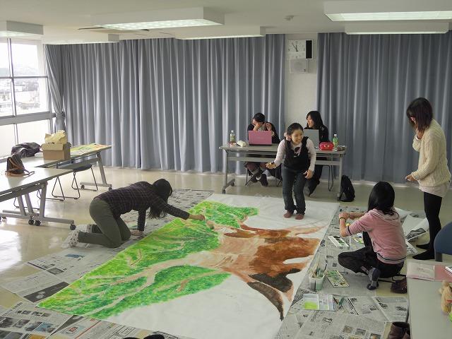 足元に広げたポスターに大きな木を描いている実行委員会の方々の写真