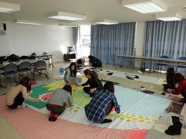 カーテンがされた白い室内で床に大きな画用紙を広げて、淳の作業をする実行委員達の写真