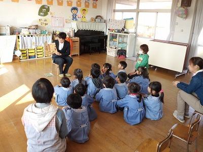 幼稚園の教室で実行員が園児たちの前で絵本の読み聞かせをしている写真
