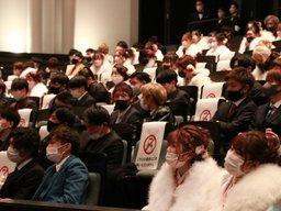 成人式の会場内の3限目の題の社会貢献活動の功績発表の結果を見る数人の成人の男女の写真