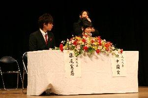 ステージ上のテーブルに着席した新成人男女二人の写真