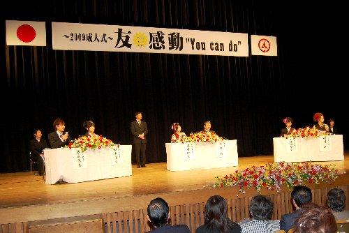 ステージ上にテーブル3個設けられ、それぞれ二人ずつ着席している新成人の写真