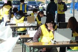 テントの下で、パソコンに向かうボランティアの女性と複数の人の写真