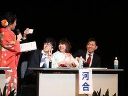 壇上の椅子に座る新成人三人が晴れ着姿の実行委員に話しかけられている写真