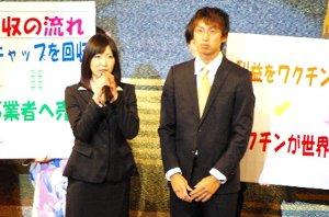 キャップ回収のプラカードの前に立つスーツの男女の写真