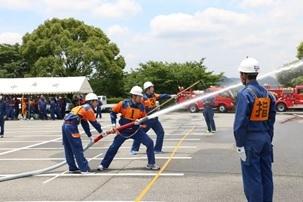 「指」と書かれたゼッケンを付けている消防隊員の奥で二人がかりで放水作業をしている消防隊員の写真