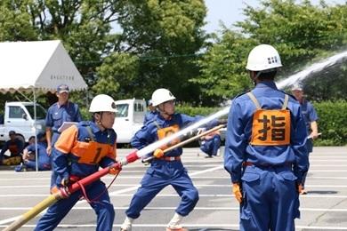 「指」と書かれたゼッケンを付けている消防隊員の奥で放水作業をしている消防隊員の写真