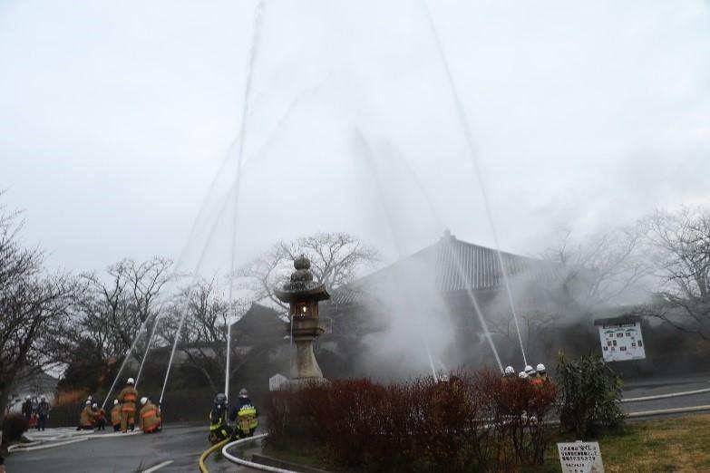 雨の中で寺院の上空に向かって放水訓練をしている消防団員たちの写真