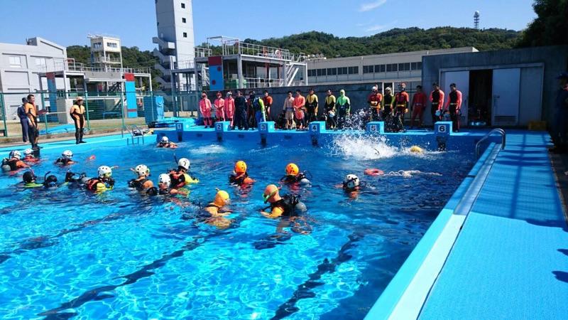 野外プールに入り水難救助の訓練をする隊員たちとプールサイドで見守る職員たちの写真