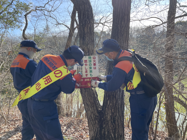 3人の紺色とオレンジ色の作業着を着て黄色の襷をかけた消防関係者が木に貼り紙をしている写真