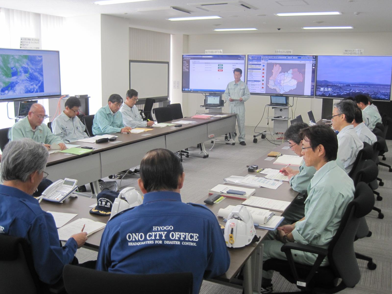 青や薄緑の作業衣を着た人々がテーブルの周りに腰掛けてモニターを見ている写真