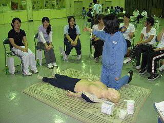 青い服を着た人が心肺蘇生人形の置かれている所で周りの椅子に座っている人に119番通報をお願いしている写真