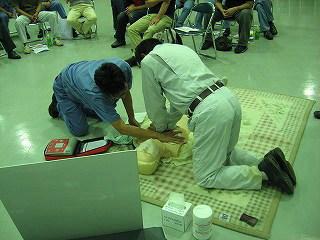青い服を着た人が心肺蘇生人形にAEDパッドを貼り付けて、同時に作業服を着た人が胸骨圧迫を行っている写真