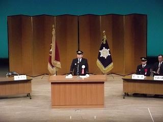 スーツを着た男性が壇上で旗を背後にして話している写真