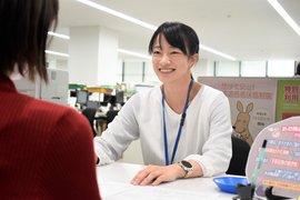 白い薄手のニットを着た女性が窓口で笑顔で対応する様子の写真