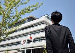 青空の下、日の丸の旗がはためいている白い建物を見上げるようにして立っているスーツ姿の男性の背中の写真