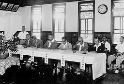 布がかけられたテーブルの前で男性6人が座っている様子の白黒写真
