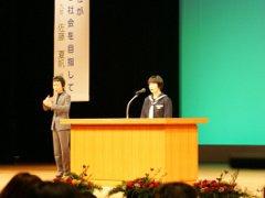 ステージ上で人権作文の発表をする女子中学生と隣で手話をする女性の写真