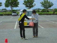 キャンペーンの黄色いタスキをかけバスケットを持った男性が駐車場で女性に啓発グッズを渡している写真