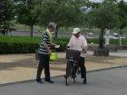 キャンペーンのたすきを掛けた人が自転車に乗った男性市民に話しかけ啓発ティッシュを渡す様子の写真