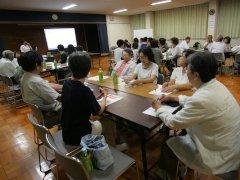 長机を合わせた複数のテーブルに分かれて座りスクリーンを見ながら話しを聞く参加者の写真
