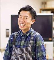青と緑のチェックのシャツとストライプ柄のエプロン姿で笑っている様子の田川雅規さんの写真