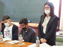 外国人の男女性が並んで席につく中、女性が立って意見を述べている写真