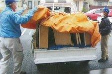 荷物を載せたトラックに二人の人がオレンジ色のシートを被せている写真