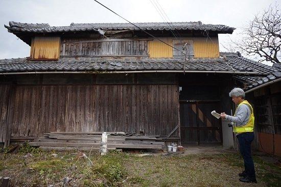 古い瓦屋根の木造住宅の下部に男性が一人立っている写真
