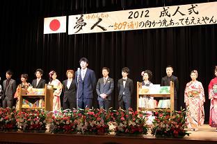 振袖やスーツを着た新成人達がステージの上に立ち、1人の男性が発言している2012年成人式の写真