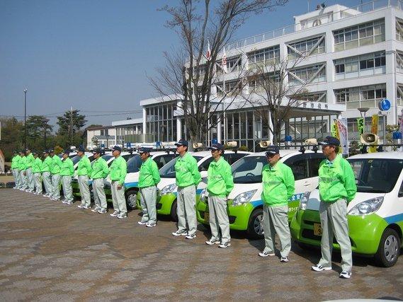 明るい緑色の上着で統一されている安全安心パトロール隊が緑と白のカラーリングの車の前で整列している様子の写真
