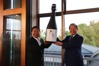 市長から倉本プロにおの恋プレミアム1年分の大きな酒瓶を贈呈している写真