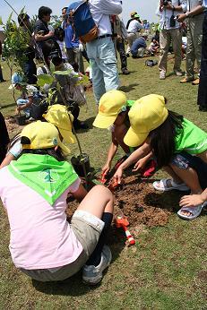 森林ボランティアの人たちが緑の少年団と一緒に芝生広場の一角に植樹をしている様子の写真