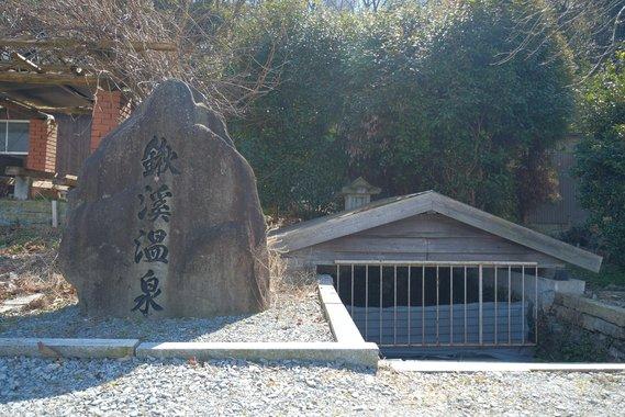 「鍬溪温泉」と書かれた石看板と、半地下になっている鍬溪温泉の入り口を正面から撮影した写真