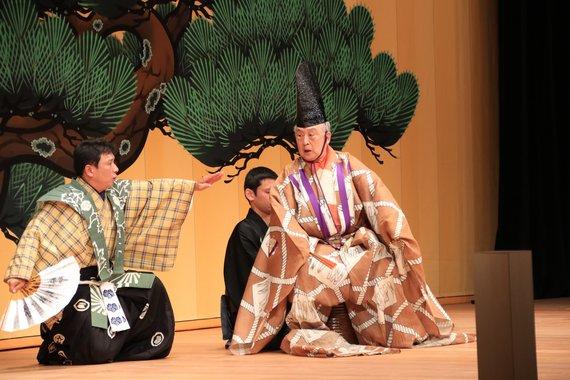 山本東次郎様がステージ上で狂言「萩大名」を演じている様子の写真
