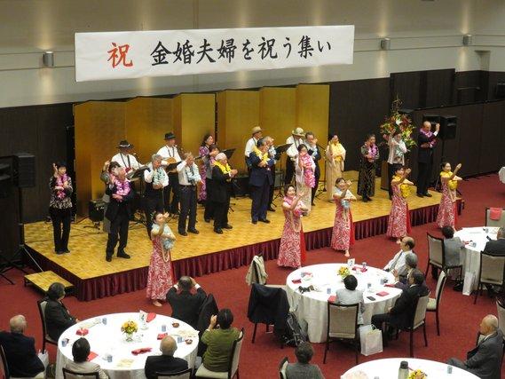 ステージの上で高齢者の方々が楽器を演奏している金婚夫婦を祝う集いの会場の様子の写真
