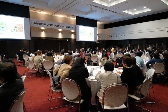 大きなスクリーンの前で沢山の人が丸テーブルに座っているお土産品の試食選考会の様子の写真