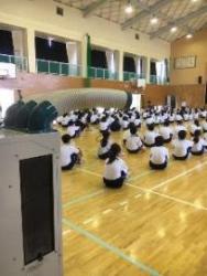 暑さ対策として体育館にスポットクーラーが設置され、そこで生徒たちが整列して体育座りをしている様子の写真