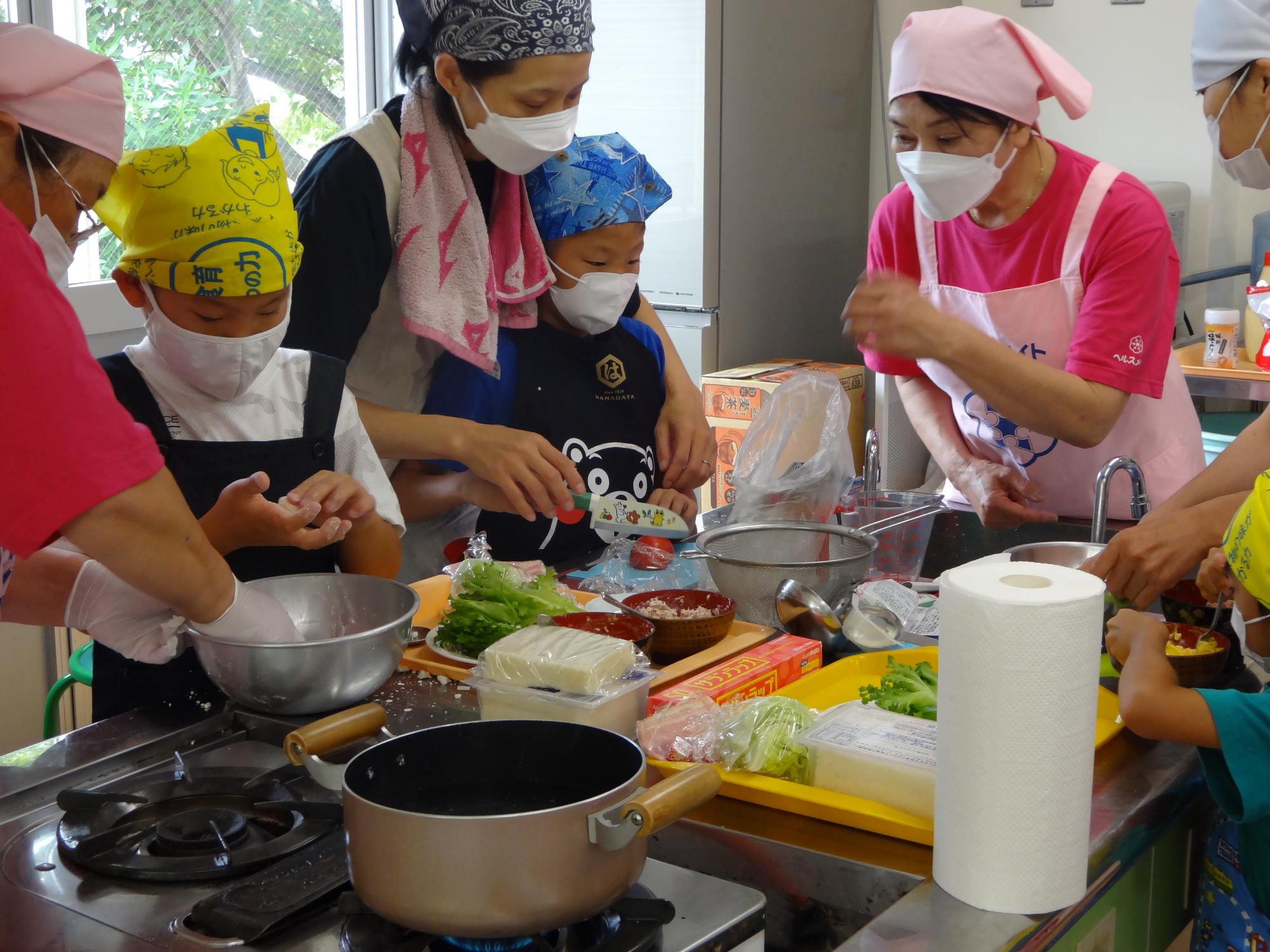 親子料理教室でテーブルの上に並べられた食材で料理を作る親子たちと補佐している職員の写真