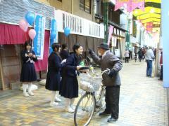 商店街で女子高生が自転車を押している男性にパンフレットを配布している写真