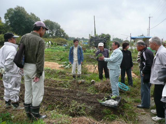 畑の中で野菜の苗を持ち説明を行う職員を複数の農園利用者が囲み熱心に話を聞く様子の写真